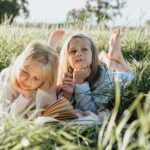 Two young girls lying on a picnic blanket in a sunny field, reading and daydreaming.